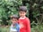 Two young children are standing together in front of a lush green background with various plants and flowers. The older child is wearing a red shirt and has their arm around the younger one, who is wearing a colorful striped sweater.