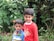 Two young children are standing together in front of a lush green background with various plants and flowers. The older child is wearing a red shirt and has their arm around the younger one, who is wearing a colorful striped sweater.