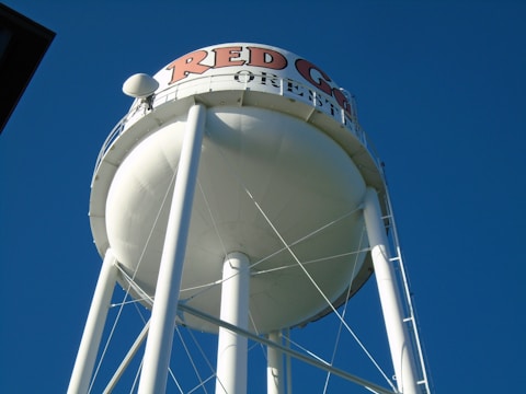A large white water tower with a round tank, supported by tall metal legs, stands prominently against a clear blue sky. The words 'RED G... OREB...' are partially visible in red and black letters at the top of the tank.