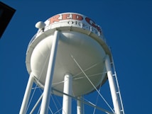 A large white water tower with a round tank, supported by tall metal legs, stands prominently against a clear blue sky. The words 'RED G... OREB...' are partially visible in red and black letters at the top of the tank.