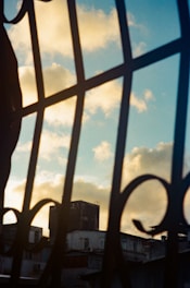 A metal fence with decorative patterns in the foreground is silhouetted against a backdrop of a blue sky with scattered fluffy clouds, partially obscuring the view of urban buildings in the distance.
