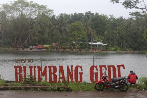 Large red letters spell out 'BLUMBANG GEDC' by the edge of a calm body of water. A small dirt path separates the letters from the water. In the foreground, a motorcycle is parked next to a person sitting on the ground. The background has lush greenery, including large trees and palm trees, with a few small structures and colorful decorations visible on the opposite shore.