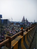 A busy urban scene featuring a crowd of people and multiple vehicles moving along a road. In the background, there is a temple with tiered roofs. The Indian flag is also visible in the scene. The foreground shows a construction barrier lined with a distinctive black-and-yellow pattern.