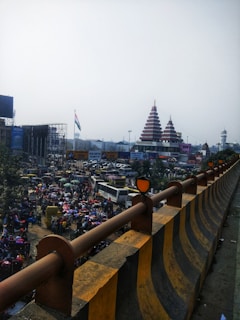 A busy urban scene featuring a crowd of people and multiple vehicles moving along a road. In the background, there is a temple with tiered roofs. The Indian flag is also visible in the scene. The foreground shows a construction barrier lined with a distinctive black-and-yellow pattern.