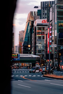 A vibrant city street with modern skyscrapers and colorful billboards. A train runs on an elevated track above the road, surrounded by a mix of cars including a taxi. The scene captures the hustle and bustle of urban life with a hint of evening illumination.