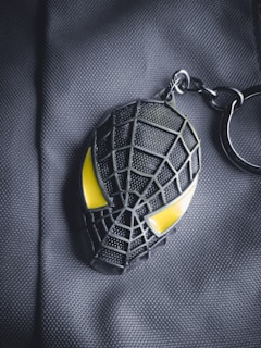 A keychain shaped like a stylized spider web face with prominent yellow eyes, laying against a textured dark gray fabric background.