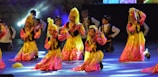Children are performing a traditional dance on stage, dressed in colorful costumes with gold and pink hues. They are holding small tambourines and appear to be in a choreographed formation with matching headpieces.