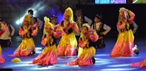 Children are performing a traditional dance on stage, dressed in colorful costumes with gold and pink hues. They are holding small tambourines and appear to be in a choreographed formation with matching headpieces.
