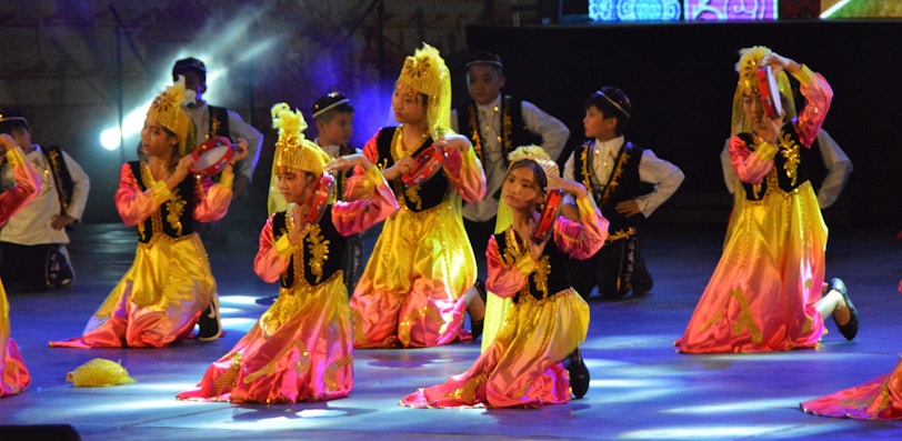 Children are performing a traditional dance on stage, dressed in colorful costumes with gold and pink hues. They are holding small tambourines and appear to be in a choreographed formation with matching headpieces.