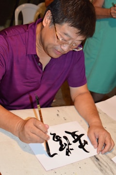 A person is focused on creating calligraphy using a brush and black ink on a piece of white paper. The individual is wearing glasses and a purple shirt, and there is another person in the background wearing a green outfit. The scene appears to be indoors, possibly at a workshop or an art class.