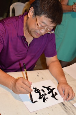 A person is focused on creating calligraphy using a brush and black ink on a piece of white paper. The individual is wearing glasses and a purple shirt, and there is another person in the background wearing a green outfit. The scene appears to be indoors, possibly at a workshop or an art class.