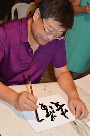 A person is focused on creating calligraphy using a brush and black ink on a piece of white paper. The individual is wearing glasses and a purple shirt, and there is another person in the background wearing a green outfit. The scene appears to be indoors, possibly at a workshop or an art class.