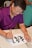 A person is focused on creating calligraphy using a brush and black ink on a piece of white paper. The individual is wearing glasses and a purple shirt, and there is another person in the background wearing a green outfit. The scene appears to be indoors, possibly at a workshop or an art class.
