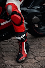 A close-up of a person wearing black and red motorcycle racing gear with branding visible, standing next to a sleek black motorcycle on cobblestone pavement.