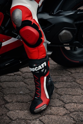 A close-up of a person wearing black and red motorcycle racing gear with branding visible, standing next to a sleek black motorcycle on cobblestone pavement.