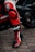 A close-up of a person wearing black and red motorcycle racing gear with branding visible, standing next to a sleek black motorcycle on cobblestone pavement.