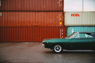 A vintage green muscle car parked on an asphalt surface with a stack of large shipping containers in the background. The containers are primarily reddish-brown with one light blue container visible. The car is partially visible, with the front side and part of the roof and windows. The sky appears overcast, creating a muted lighting effect.