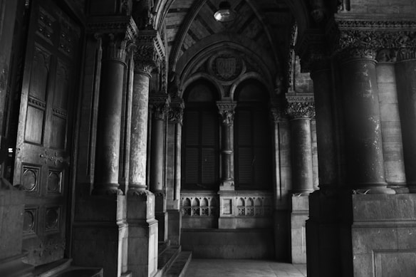 A black and white interior image depicting a historical architectural space with ornate stone columns and intricate carvings. There are large archways and decorative details lining the walls. Light subtly illuminates sections of the stonework, highlighting its texture.