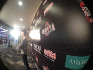 A person in a striped shirt stands with arms crossed in front of a promotional backdrop adorned with various brand logos. The area is well-lit with overhead lights and a bright light source on the side. The setting appears to be an event space with other booths or setups visible in the background.