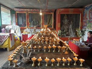 A richly decorated room with an altar displaying numerous lit butter lamps arranged in rows. The background features Tibetan Buddhist art and statues enclosed in ornate cabinets. A person is seated to the right, engaged in reading or writing, surrounded by more religious items and offerings.
