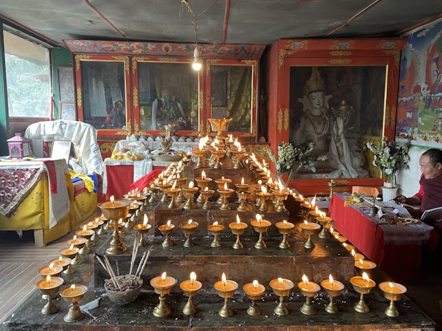 A richly decorated room with an altar displaying numerous lit butter lamps arranged in rows. The background features Tibetan Buddhist art and statues enclosed in ornate cabinets. A person is seated to the right, engaged in reading or writing, surrounded by more religious items and offerings.