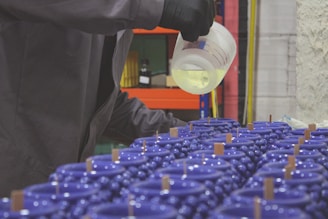 A person wearing black gloves is pouring a liquid from a measuring cup into a series of multiple blue containers, which appear to be candles with wooden wicks. The setting resembles a workshop or factory space.