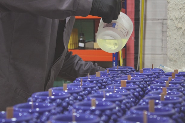 A person wearing black gloves is pouring a liquid from a measuring cup into a series of multiple blue containers, which appear to be candles with wooden wicks. The setting resembles a workshop or factory space.