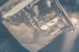 A close-up view of a glass container, possibly a beaker, showing small white crystalline particles adhered to its interior surface. The background is blurred, with a darker and slightly out-of-focus setting.