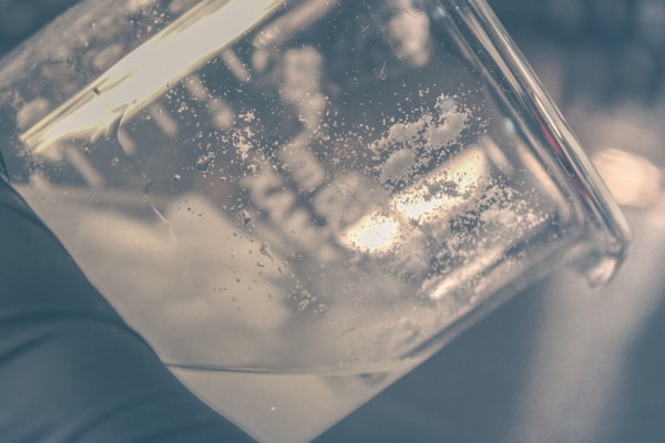 A close-up view of a glass container, possibly a beaker, showing small white crystalline particles adhered to its interior surface. The background is blurred, with a darker and slightly out-of-focus setting.