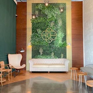 A stylish indoor space with a vibrantly green vertical garden wall, adorned with lush plants and geometric metal wall art. Below the wall is a white tufted sofa, and adjacent to it, a modern lamp. In the foreground, there are chic chairs and small wooden tables on a polished concrete floor.