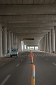 An empty road under a large concrete structure with tall columns. Orange traffic cones are lined along the middle, marking the lane. A small white vehicle is parked on the left side. The scene appears to be deserted with a distant view of greenery visible beyond the structure.