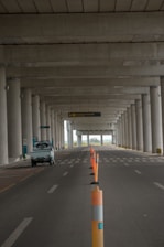 An empty road under a large concrete structure with tall columns. Orange traffic cones are lined along the middle, marking the lane. A small white vehicle is parked on the left side. The scene appears to be deserted with a distant view of greenery visible beyond the structure.