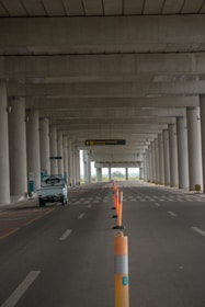 An empty road under a large concrete structure with tall columns. Orange traffic cones are lined along the middle, marking the lane. A small white vehicle is parked on the left side. The scene appears to be deserted with a distant view of greenery visible beyond the structure.