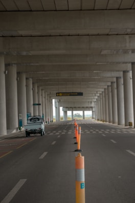 An empty road under a large concrete structure with tall columns. Orange traffic cones are lined along the middle, marking the lane. A small white vehicle is parked on the left side. The scene appears to be deserted with a distant view of greenery visible beyond the structure.