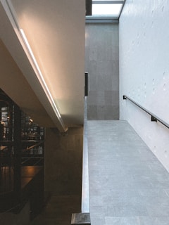 A modern interior space featuring a split-level design with a sloping ramp on the right side. The space is divided by a wall, with one side illuminated by a linear light fixture and the other side darker with visible structural elements. The surfaces are sleek and minimalistic, with tiles on the ramp.
