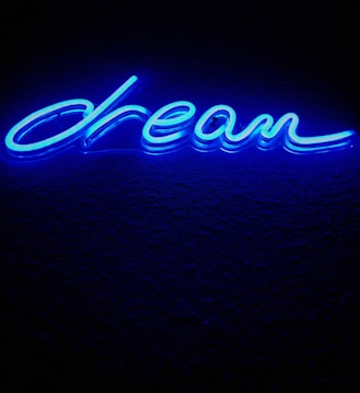 A bright blue neon sign displaying the word 'dream' written in cursive. The sign emits a glowing light against a dark background, creating a vibrant contrast.