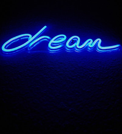 A bright blue neon sign displaying the word 'dream' written in cursive. The sign emits a glowing light against a dark background, creating a vibrant contrast.