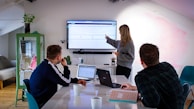 Three people are in an office setting, gathered around a table with laptops and notebooks. One person is standing and presenting information on a large screen mounted on the wall, pointing to graphs while the other two are seated and attentively listening. The room is well-lit and decorated with a plant and shelves.