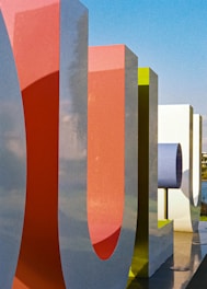 Large, colorful, stylized letters with a smooth finish stand upright, casting shadows on the ground. The letters are three-dimensional and painted in vibrant shades of red, green, and white against a bright blue sky.