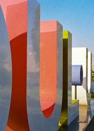Large, colorful, stylized letters with a smooth finish stand upright, casting shadows on the ground. The letters are three-dimensional and painted in vibrant shades of red, green, and white against a bright blue sky.