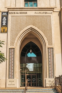 A grand entrance featuring an arched doorway framed with intricate geometric patterns and colorful mosaic tiles. The top section displays 'Souk Al Bahar' signage with Arabic script and a promotional banner on the left side. A hint of greenery is visible beside the steps leading up to the door.
