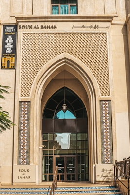 A grand entrance featuring an arched doorway framed with intricate geometric patterns and colorful mosaic tiles. The top section displays 'Souk Al Bahar' signage with Arabic script and a promotional banner on the left side. A hint of greenery is visible beside the steps leading up to the door.