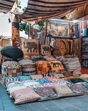 A market stall showcasing a variety of colorful, patterned textiles. There are numerous pillows, cushion covers, and decorative throws stacked and arranged neatly. The textiles display intricate designs, including elephants and mandalas. Some items are hung up on the walls while others are laid out on the ground. The stall is set up outdoors, and a striped cover provides shade.