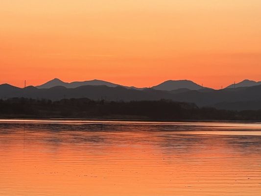 The image features a serene landscape with a calm body of water in the foreground reflecting the vibrant hues of an orange sunset. Silhouetted mountains line the horizon, creating a striking contrast against the brightly colored sky.