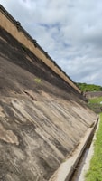 A large, sloped concrete structure resembling a dam or retaining wall stretches into the distance under an overcast sky. Sparse vegetation grows on sections of the structure, and a narrow drainage channel runs alongside. Green foliage covers the hillside visible in the background.