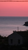 A tranquil seaside view at sunset with a silhouetted cottage in the foreground. The sky displays a gradient of pink and orange hues, while the calm sea reflects these colors. A few small boats can be seen in the distance, and the silhouette of trees is visible against the horizon.