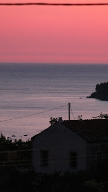 A tranquil seaside view at sunset with a silhouetted cottage in the foreground. The sky displays a gradient of pink and orange hues, while the calm sea reflects these colors. A few small boats can be seen in the distance, and the silhouette of trees is visible against the horizon.
