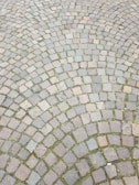 A pattern of cobblestone paving arranged in a circular design, with varying shades of gray and brown stones. Small patches of green moss are visible between some of the stones, adding texture.