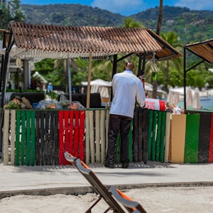 A person stands at a colorful wooden market stall on a sandy beach. The stall displays various fruits and vegetables under a shaded awning. Nearby, deck chairs are positioned facing the sea, suggesting a relaxed beach setting with lush greenery and mountains in the background.