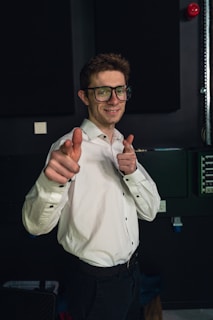 A person wearing glasses and a white shirt points both index fingers forward in a playful gesture. The background appears to be a dark room with some electrical equipment visible.